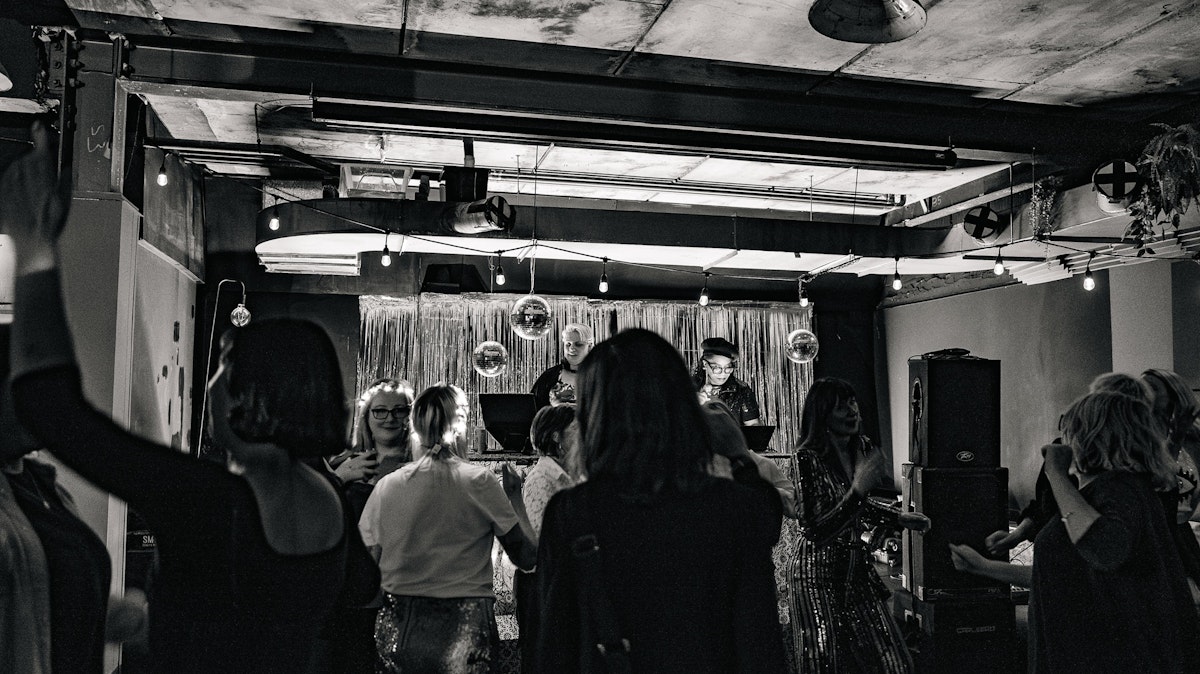 Crowd of people dancing in front of two DJs at a Grief Disco, with disco balls and silver streamers overhead